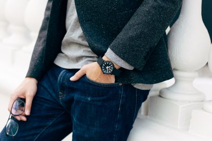 Man resting against a wall with his hand in his pocket, revealing a black &amp; white watch on wrist