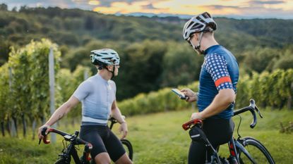 Two riders stand in front of a beautiful vineyard view, one checks his phone