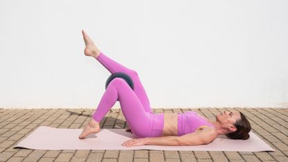 A woman practices Pilates toe taps on a mat outdoors. She is lying on the mat with her arms by her side and her knees bent; there is a Pilates ball between her knees. One foot taps the floor while the other is elevated. Behind her is a white-washed wall.