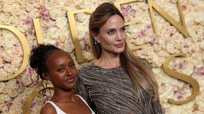 Angelina Jolie in a glittery black gown and her daughter Zahara standing in front of a rose covered Golden Globes step and repeat