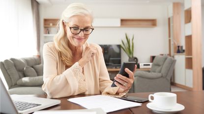 An older woman smiles as she uses a calculator and looks at paperwork in her living room.