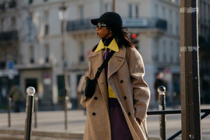 Attendee at paris fashion week menswear fall winter 2023 wearing camel coat, yellow shirt, crossbody bag, black hat, and sunglasses 