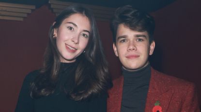 Samuel Chatto wears a red jacket and black top as he poses with girlfriend Eleanor Ekserdjian