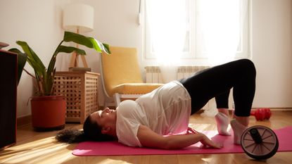 A woman performs a glute bridge at home. She is on a yoga mat, with her toes and upper back resting on the mat and hips elevated. Behind her we see a chair, window, table, lamp and plant. Next to her is an abs wheel.