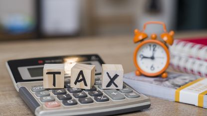 Wooden blocks spell out the word tax on top of a calculator next to an alarm clock sitting on a stack of cash.