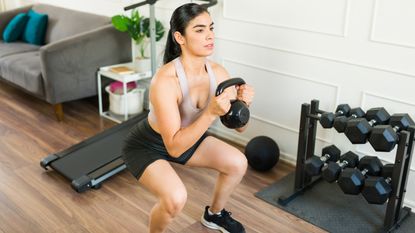 A woman holds a kettlebell close to her chest and performs a squat. Her knees are bent and feet hip-width apart. Behind her is a couch, treadmill, and dumbbell rack.