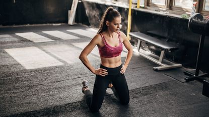 Woman wearing sportswear kneels on gym floor with her hands on her hips