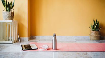 Yoga mat on tile floor with laptop, water bottle and headphones to represent a home gym set up