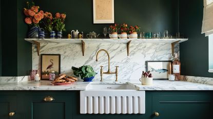 dark green kitchen with marble countertops and backsplash, fluted white sink, brass hardware, marble shelf with flowers, glassware, artwork