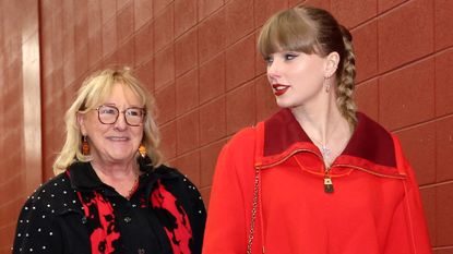 Taylor Swift wearing a red pullover walking next to Donna Kelce next to a red brick wall