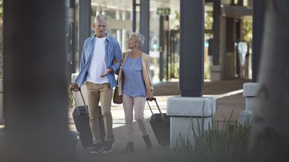 An older couple walk outside of an airport, pulling their luggage. 