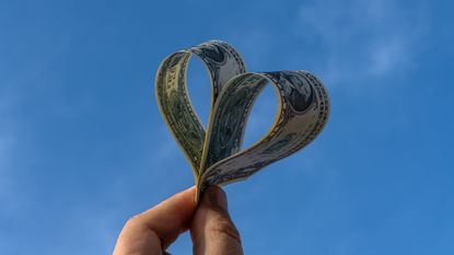 Two dollar bills shaped into a heart held against the backdrop of a blue sky.