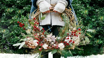 A woman showcasing festive winter wreath ideas, standing in the garden on a patch of snow., holding a foliage and berry wreath.