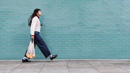 Young woman with mesh bag on footpath by turquoise brick wall 