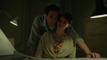 adrien brody leans over felicity jones as they look over blue prints in a still from the brutalist