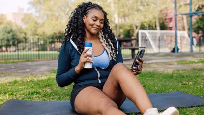 A woman relaxes on an exercise mat outside and looks at her phone. She is seated on the mat, with one leg stretched out and the other bent. She holds a water bottle in one hand and her phone in the other. Behind her we see a park, with black iron fencing and trees.