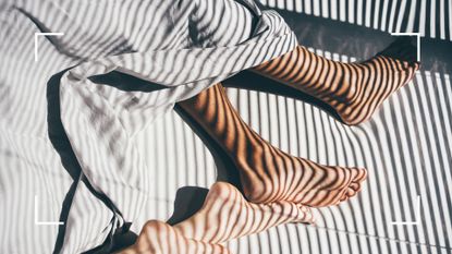 Bird&#039;s eye view of couples feet in bed during hot morning with sun coming through slatted blinds, representing how to sleep in the heat