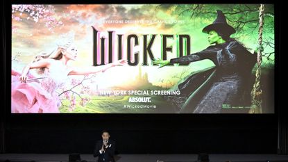 screening of Wicked movie, with the title still showing on a big screen and director Jon Chu speaking into a microphone