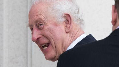 A headshot of King Charles smiling and looking to the side wearing a black coat