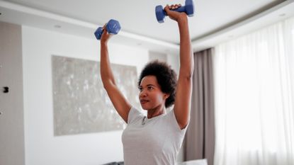 woman wearing a white tshirt in a white living room setting holding two blue small dumbbells with arms extended overhead. 