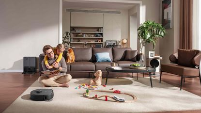 Man sitting on cream carpet with young blond child on his back, toys and a robot vacuum in front