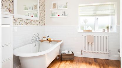 White freestanding bath in a white bathroom with floral wallpaper behind it and wooden floor