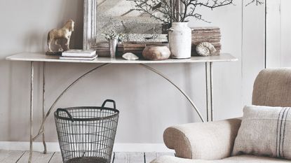 A living room in a neutral colour scheme with a faded mirror on a weathered console table with a beige linen chair in front