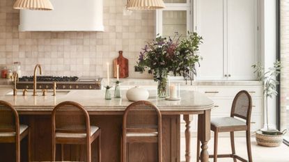 A neutral kitchen with a large wooden and marble island that has four wooden bar chairs around it. The backsplash is made of pink-beige tile and there is white cabinetry and a white hutch