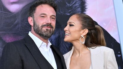 Ben Affleck wearing a black suit jacket and white shirt standing next to Jennifer Lopez who is smiling at him