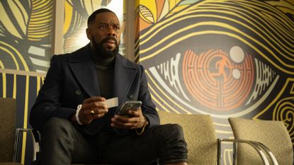 Colman Domingo as Muncie Daniels, holding a business card and a phone while sitting in front of a wall with a mural of an eye-shaped maze, in Episode 101 of The Madness.