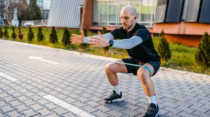 A man in sportswear performs a banded lateral walk outside. He has a short looped resistance band wrapped around his thighs and he is squatting down, with knees bent and thighs parallel to the floor. He holds his arms straight out in front of him. Behind him we see a building with large windows and a line of young fir trees.