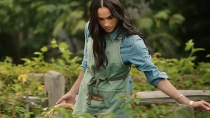 Meghan Markle wearing an apron and denim shirt walking through a garden and holding a basket