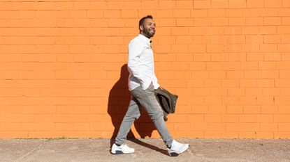 A man walks along a street smiling, carrying his jacket in his hand. He wears jeans, a shirt and sneakers and behind him is a painted orange wall.