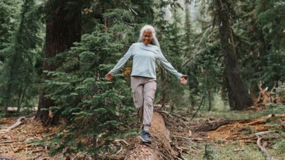 A woman walks along a fallen tree in a forest, smiling and holding her arms out to her sides. We see several large trees behind her.