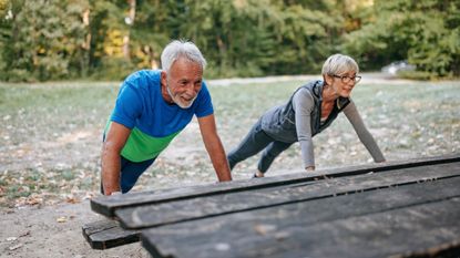 A man and a woman in sportswear are performing elevated push-ups on a picnic table outdoors. Their arms are straight and they are leaning over the table, holding themselves at an angle. Behind them we see grass and bushes.