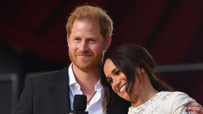 Prince Harry wears a suit jacket and an open white shirt as he hugs wife Meghan Markle at Global Citizen, she is wearing a white dress adorned with silver detailing