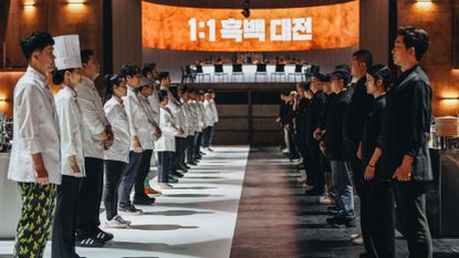 Two rows of chefs (the White Spoons, left, and the Black Spoons, right) stand facing each other, with a circular dining table in the back and a screen reading &quot;1:1&quot; with a Korean phrase, in &#039;Culinary Class Wars.&#039;