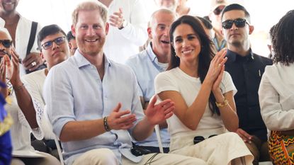 Prince harry and Meghan Markle sitting in a row of chairs clapping and smiling 