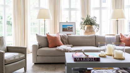 living room with cream drapes, gray couch, gray leather armchair, coffee table with books and candles, rust couch pillows, floor lamps,