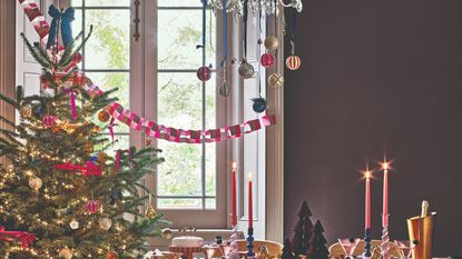 A dining room with the table set for Christmas and the room decorated with paper garlands, baubles hanging from a chandelier and a tree with a bow for a tree topper