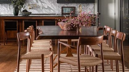 Image of a wooden dining table with 10 wooden dining chairs around it. There is a beige/brown stripped rug under the table and a bowl of pink flowers on top.