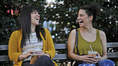 abbi and ilana sit on a park bench laughing in broad city