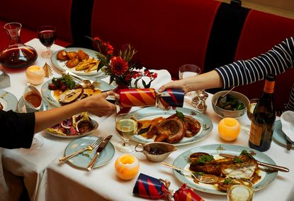 Two people hold a Christmas cracker on top of a festive dining table filled with food, drinks, and candles.