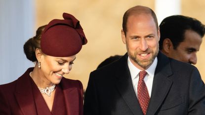 Kate Middleton wears a maroon coat dress and matching hat and smiles while husband Prince William puts his arm around her