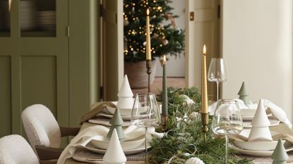 A neutral dining toom with a long table decorated with simpler ceramic trees and candlesticks, lit. A christmas tree visible through doors behind. 