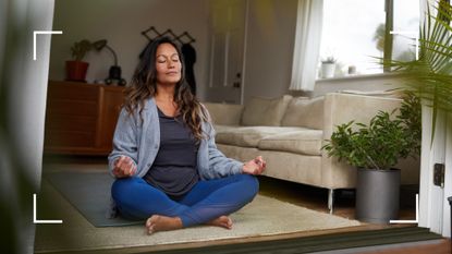 Woman sitting with legs crossed in the middle of her living room between plants, representing yoga over 50