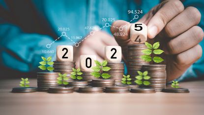 A man&#039;s hand flipping a wooden block cube from 2024 to 2025. Wooden block cubes are stacked on top of coins with plant shoots growing out of them. 