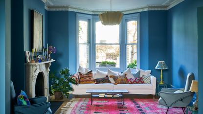 A blue living room with statement pink rug, large bay window, mantelpiece and white sofa with colourful cushions