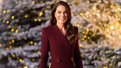 Kate Middleton wearing a burgundy coat and smiling in front of lit up Christmas trees