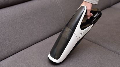 A handheld vacuum being used on a gray fabric sofa